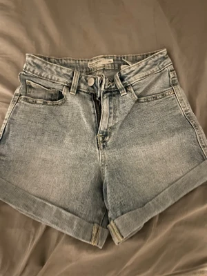 Ljusblå denimshorts från H&M - Snygga ljusblå denimshorts från H&M med hög midja och klassisk femficksdesign. Shortsen har uppvikta benslut och stängs med dragkedja och knapp. Perfekta för varma dagar och enkel att matcha med allt.