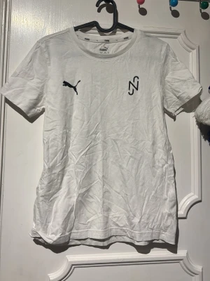 Vit Puma t-shirt med färgglad rygg - Vit t-shirt från Puma med svart logga och grafiskt tryck framtill. Baksidan har ett stort färgglatt motiv med taggiga former i blått, rött, gult och lila. Klassisk passform och rund halsringning. Perfekt för dig som gillar sportig och unik stil.