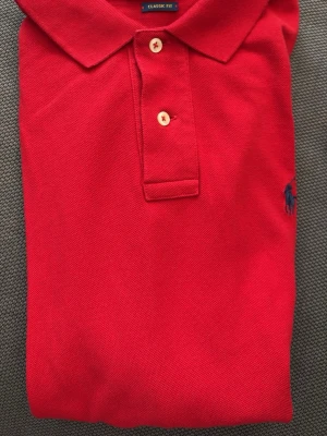 Röd Piké från Ralph Lauren - Klassisk röd pikétröja från Polo Ralph Lauren med krage, två knappar och broderad logga på bröstet. Tröjan har en tidlös design och är perfekt för en stilren look. Storlek Medium. Snabb Leverans!