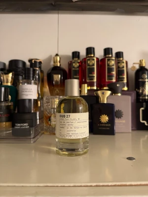 Le Labo Oud 27 Eau de Parfum - Le Labo Oud 27 är en exklusiv parfym i en stilren, genomskinlig glasflaska med metallock. Doften är känd för sina djupa, träiga och rökiga oud-noter. Flaskan har en minimalistisk etikett med svart text och rymmer 50 ml.