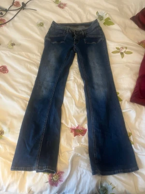 Mörkblå bootcut jeans - Säljer ett par mörkblå bootcut jeans med klassisk femficksdesign och snygga slitningar framtill. Jeansen har låg midja och vida ben, perfekta för en avslappnad och trendig look. Tillverkade i denim med kontrastsömmar. Midja är 33 cm rakt över och inner benlängd är 72 cm