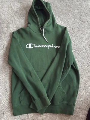 Grön Champion hoodie med logga - Snygg grön hoodie från Champion med stor vit logga över bröstet. Klassisk modell med huva och vita dragsnören samt känguruficka framtill. Perfekt för en avslappnad och sportig stil.