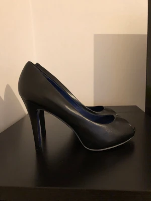 Svarta peep-toe pumps med hög klack - Säljer ett par stilrena svarta pumps med peep-toe och hög klack. Skorna har en elegant siluett och blå insida. Äkta läder. Perfekta för dig som vill ha en klassisk men modern look.