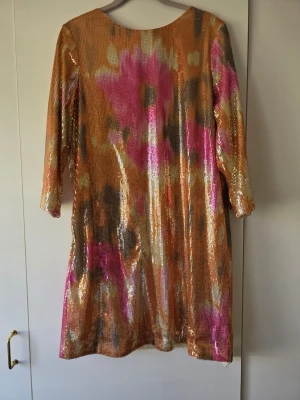 Glittrig festklänning med paljetter - Säljer en glittrig festklänning med paljetter i orange, rosa och guld. Klänningen har ett abstrakt mönster, trekvartsärm och rund halsringning. 