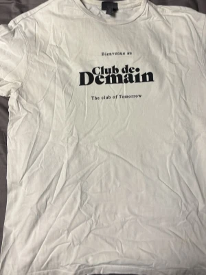 Vit t-shirt Club de Demain print - Vit t-shirt med svart textprint 'Club de Demain' och 'The club of Tomorrow' på bröstet. Klassisk rund hals och korta ärmar. Perfekt för en avslappnad och trendig look.