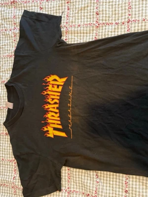 Svart Thrasher t-shirt med flames - Säljer en svart t-shirt från Thrasher med den ikoniska loggan i orange och gula flames på bröstet. T-shirten är i bomull och har en klassisk passform med korta ärmar. 
