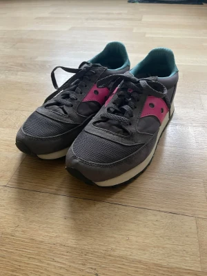 Saucony grå/rosa/blå sneakers - Snygga Saucony sneakers med rosa och blå detaljer på sidorna. Skorna har en vit och svart sula. Perfekta för dig som gillar en sportig och retro stil.