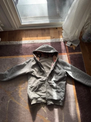 Grå zip hoodie från Burberry - Säljer en grå hoodie från Burberry med klassiskt rutigt foder i huvan. Dragkedjan funktionerar inte som den ska. Hoodien har fickor framtill och justerbar dragsko i huvan. Perfekt för dig som gillar stilrena plagg med en twist av lyx.