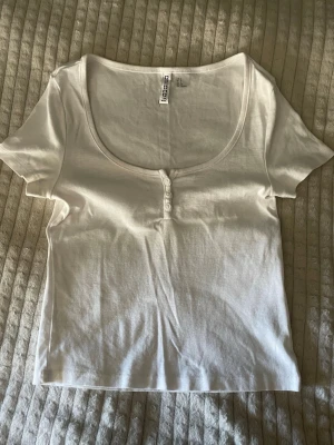Vit kortärmad topp från H&M Divided - Vit kortärmad topp från H&M Divided med rund, djup ringning och tre små knappar framtill. Topp i mjuk bomull som passar perfekt till jeans eller kjol. Enkel och stilren design som är lätt att matcha. Inga defekter och använd vid tre tillfällen.