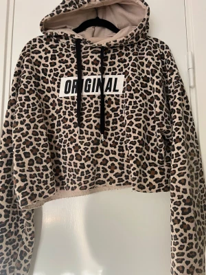 Leopardmönstrad cropped hoodie - Säljer en leopardmönstrad cropped hoodie med svartvita snören och trycket 'ORIGINAL' på bröstet. Hoodien har huva, långa ärmar och ribbade muddar. Insidan av huvan är beige och materialet känns mjukt och bekvämt.