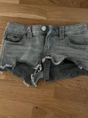Gråa jeansshorts med fransar - Säljer ett par gråa jeansshorts med slitna detaljer och fransig kant. Shortsen har klassisk femficksdesign, knappstängning och låg midja. Perfekta för en avslappnad och cool stil.