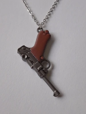 Halsband med pistolhänge i metall - Unikt halsband med ett hänge, en pistol. Hänget är i silverfärgad metall med ett brunt handtag och detaljerad design. Kedjan är tunn och silverfärgad, perfekt för dig som gillar statement-accessoarer med edge. Materialet rostfritt stål. 