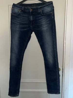 Mörkblå slim fit jeans från Nudie - Snygga mörkblå slim fit jeans från Nudie Jeans med klassisk femficksmodell och subtila slitningar framtill. Jeansen har en skön stretch och är tillverkade i denim med kontrastsömmar och läderpatch bak i midjan.