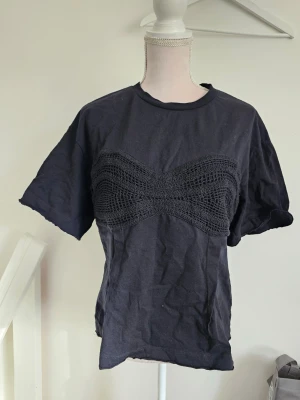 Svart t-shirt med virkad detalj - Cool svart t-shirt med unik virkad detalj över bröstet. T-shirten har rund halsringning och korta ärmar. Perfekt för dig som vill ha en basic tee med en twist.