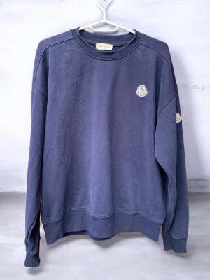 Mörkblå sweatshirt från Moncler - En stilren mörkblå sweatshirt från Moncler med rund halsringning och ribbade muddar. Tröjan har Monclers klassiska logotyp på bröstet och ärmen. Tillverkad i mjukt bomullsmaterial som är skönt mot huden. Perfekt för en avslappnad och trendig look.