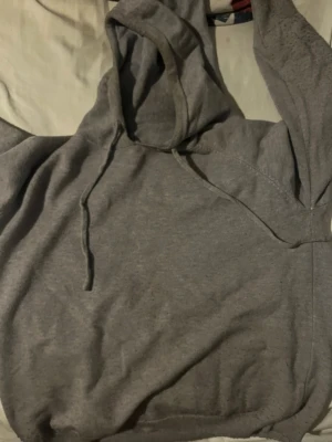 Grå basic hoodie med huva - En enkel grå hoodie med huva och dragsko. Mjuk och skön, perfekt för chill dagar eller när du vill ha en avslappnad stil. Tillverkad i bomullsblandning för extra komfort. Klassisk passform utan tryck eller logga. Tröjan har en fläck därav priset. Se bild 2