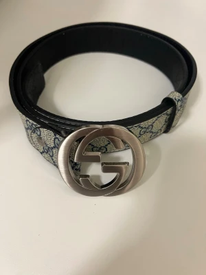 Gucci bälte med GG-spänne - Snyggt Gucci bälte med klassiskt GG-spänne i silverfärgad metall. Bältet har ett mönstrat tygband i beige och blått med Guccis ikoniska logga och insida i svart skinn. Perfekt accessoar för att lyfta din outfit.