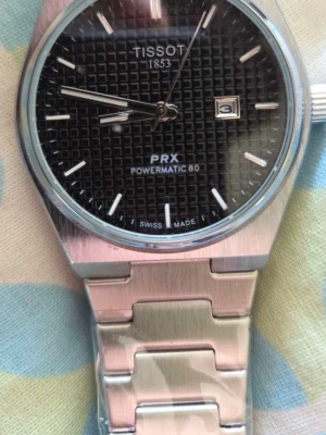Tissot PRX Powermatic 80 klocka - Snygg Tissot PRX Powermatic 80 klocka med svart urtavla och datumfönster. Klockan har ett stilrent rutmönster på tavlan och ett armband i silverfärgad metall. Perfekt accessoar för dig som gillar klassisk och modern design.