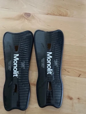  Monolit - Säljer ett par svarta benskydd från Monolit med kolfibermönster. De har en ergonomisk form och ventilationshål för extra komfort. Perfekta för fotboll eller annan kontaktsport där skydd behövs.