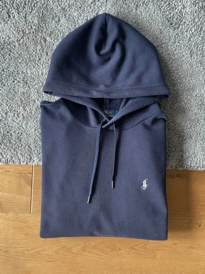 Mörkblå hoodie från Polo Ralph Lauren - Snygg mörkblå hoodie från Polo Ralph Lauren med klassisk huva och dragsko. Tröjan har en broderad vit logga på bröstet och en stor magficka framtill. Perfekt för en avslappnad och stilren look. Tillverkad i mjukt bomullsmaterial.