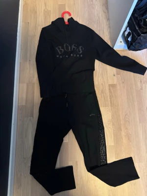 Svart hoodie från Hugo Boss - Snygg svart hoodie från Hugo Boss med stor logga framtill i silverprickar. Klassisk modell med huva och dragsko samt känguruficka. Perfekt för en stilren och avslappnad look. Materialet är mjukt och bekvämt, passar perfekt till vardags eller när du vill chilla.