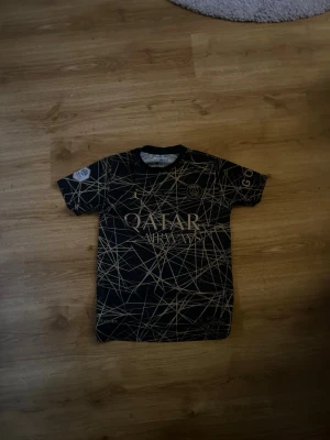 PSG Messi matchtröja svart/guld - Snygg svart PSG matchtröja med guldiga detaljer och tryck. Framsidan har Qatar Airways-logga och PSG-emblem, baksidan har Messi och nummer 30. Unik design med abstrakta guldlinjer över hela tröjan. Perfekt för fotbollsfans som vill sticka ut.