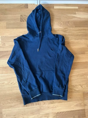 Mörkblå hoodie från Fred Perry - Mörkblå hoodie från Fred Perry med broderad logga på bröstet. Perfekt för en avslappnad och stilren look. Hör av er vid funderingar eller fler bilder!