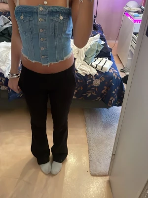 Ljusblå denim korsett-topp med knappar - Trendig ljusblå korsett-topp i denim med knappar framtill och rå kant nertill. Toppen är axelbandslös och har en croppad passform som ger en edgy vibe. Perfekt för dig som vill sticka ut med en cool och unik stil.