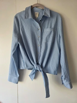 Blåvit randig knytblus från ONLY - Snygg blåvit randig blus från ONLY med knytning framtill och klassisk krage. Blusen har långa ärmar med manschett och tryckknappar framtill. Skön varma dagar.