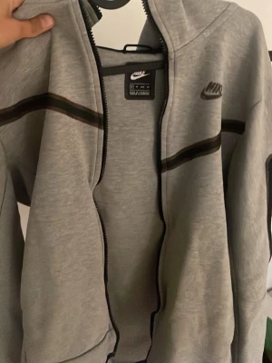 Grå zip hoodie från Nike - Snygg grå hoodie från Nike med dragkedja och svart Nike-logga på bröstet. Hoodien har svarta och bruna ränder över bröstet och armarna samt en klassisk huva. Tillverkad i mjukt bomullsmaterial som är skönt att ha på sig.Sändrig zip går ej att knäppa. Pris kan diskuteras!