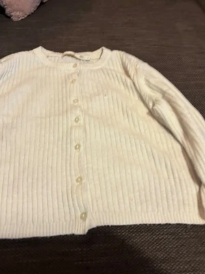 Vit ribbad tröja med knappar - Snygg vit ribbstickad kofta med rund hals och knäppning framtill. Koftan har långa ärmar och är tillverkad i ett mjukt material som känns skönt mot huden. Perfekt att slänga över en t-shirt eller linne för en chill look.