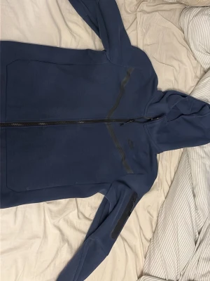 Nike tech marinblå - Snygg blå hoodie från Nike med hel dragkedja framtill och huva. Hoodien har svarta detaljer längs bröstet och en ficka med dragkedja på ärmen. Perfekt för en sportig och avslappnad stil.