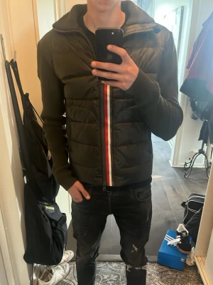 Mörkgrön cardigan från Moncler - Säljer en mörkgrön puffercardigan med stickade ärmar och huva. Jackan har en dragkedja framtill med röd, vit och blå rand längs öppningen. Två sidofickor med dragkedja och ribbade muddar vid ärmslut. Perfekt för sena sommarkvällar, kalla vinterdagar, höst, vår