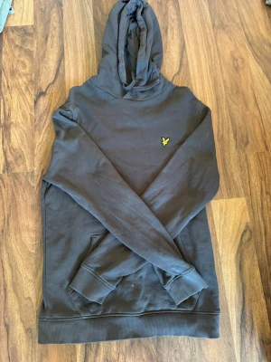 Mörkgrå hoodie från Lyle & Scott - Snygg mörkgrå hoodie från Lyle & Scott med klassisk gul logga på bröstet. Hoodien har huva med dragsko och en stor magficka framtill. Tillverkad i mjukt bomullsmaterial och har ribbade muddar vid ärmslut och nederkant.