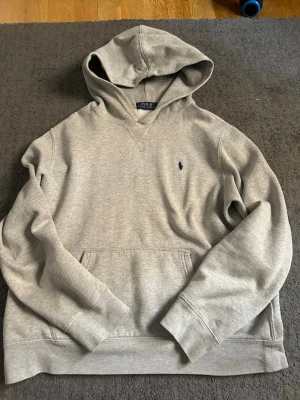 Grå hoodie från Polo Ralph Lauren - Snygg grå hoodie från Polo Ralph Lauren med klassisk broderad logga på bröstet. Hoodien har känguruficka, ribbade muddar och mjukt material som känns skönt mot huden. Perfekt för en avslappnad och stilren look.