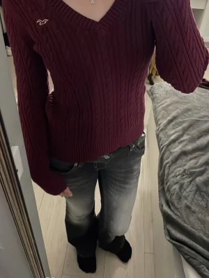 Vinröd kabelstickad tröja Hollister - Vinröd kabelstickad tröja från Hollister med V-ringning och lång ärm. Tröjan har ribbade muddar och nederkant samt en liten vit logga broderad på bröstet. Perfekt för lager-på-lager och snygg till jeans.