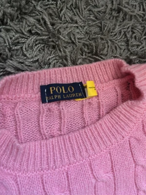 Rosa sweater Polo Ralph Lauren - Rosa kabelstickad tröja från Polo Ralph Lauren med rund halsringning och den klassiska broderade loggan på bröstet. Tröjan är stickad i ett mjukt material och har ribbade muddar vid hals, ärmslut och nederkant. Perfekt för dig som gillar preppy och tidlös stil. passar till båda män och tjej 