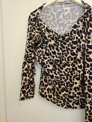 Leopardmönstrad blus med drapering - Trendig blus med leopardmönster i beige, svart och brunt. Blusen har en snygg draperad ringning och långa ärmar. Tillverkad i ett mjukt, lätt tyg som ger ett snyggt fall. Perfekt för dig som vill sticka ut med ett djurmönster.