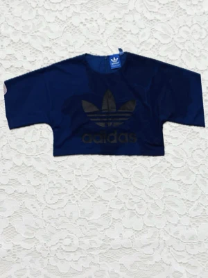Mörkblå croppad topp från Adidas - Croppad mörkblå topp från Adidas med stort svart logotryck framtill. Toppen har vida, korta ärmar och är tillverkad i polyester. Perfekt för en sportig och trendig look.