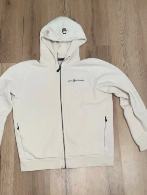 Vit Sail Racing Zip-Hoodie - Snygg vit hoodie från Sail Racing med dragkedja framtill och två fickor med svarta detaljer. Broderad logga på bröstet och en liten logga på huvan. Mjuk bomullskänsla och klassisk passform, perfekt till jeans eller joggers.