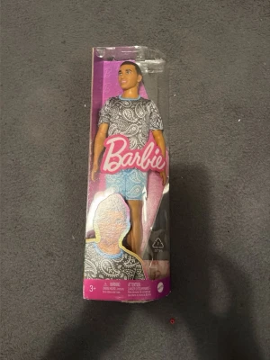 Barbie Ken docka - Säljer en Ken-docka från Barbie-serien. Dockan har en svartvit paisleymönstrad t-shirt och blå shorts. Förpackningen är rosa och transparent med Barbie-loggan framtill. Perfekt för samlare eller som present till någon som gillar Barbie.