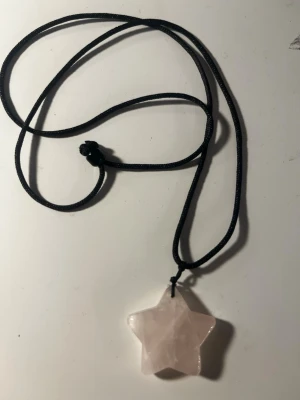 Halsband med stjärnformad rosa sten - Ett halsband med svart snöre och en stjärnformad berlock i ljusrosa sten, troligen rosenkvarts. Enkelt och stilrent smycke som passar till många olika looks. Perfekt för dig som gillar unika detaljer och kristaller.