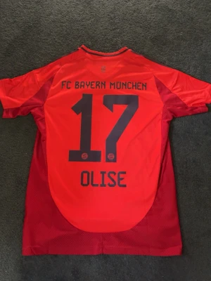Röd FC Bayern München matchtröja - Säljer en röd FC Bayern München matchtröja från Adidas med nummer 17 och namnet Olise på ryggen. Tröjan har svarta detaljer, klubbmärke och sponsorlogga framtill samt klassiska Adidas-ränder på axlarna. Perfekt för fotbollsfans!