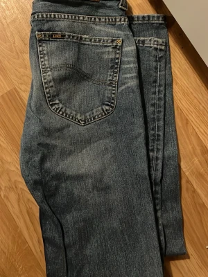 Blå raka jeans från Lee - Klassiska blå jeans från Lee med rak passform och femficksmodell. Jeansen har en snygg tvättad look och detaljer i metall på knappar och nitar. Tillverkade i slitstarkt denimtyg som ger en tidlös och avslappnad stil.