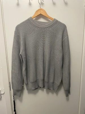 Ljusgrå stickad tröja unisex - En stilren ljusgrå stickad tröja med rund halsringning och ribbade muddar vid ärmslut och nederkant. Tröjan har en rak passform och är tillverkad i ett mjukt, grovt stickat material som passar perfekt för lager på lager.