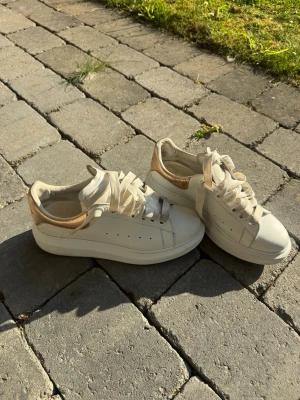 Vita Alexander McQueen sneakers - Stilrena vita sneakers från Alexander McQueen med metallicguld-detalj på hälen och klassiska breda snören. Skorna har en chunky sula och är tillverkade i läder med perforerade sidor för extra ventilation. Perfekta för dig som gillar en clean och lyxig look.