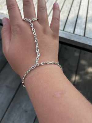 Silvrigt handkedjearmband - Snyggt handkedjearmband i silverfärgad metall som kopplar ihop handled och finger med en kedja. Armbandet har en enkel, stilren design med ovala länkar och ett dekorativt lås. Perfekt för dig som vill ha något unikt och trendigt till din outfit.        Skriv om du har frågor.☺️