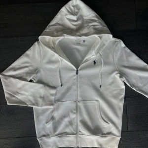 Vit hoodie från Polo Ralph Lauren - Säljer en fräsch vit hoodie från Polo Ralph Lauren med dragkedja och huva. Tröjan har ett mörkblått broderat logomärke på bröstet, snörning vid huvan och två stora fickor framtill. Tillverkad i mjuk bomull, perfekt för chill dagar.
