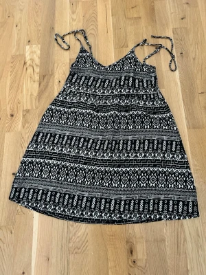 Svartvit mönstrad klänning H&M Divided - Kort svartvit klänning från H&M Divided med grafiskt mönster och tunna axelband som knyts. Klänningen har en avslappnad passform och är perfekt för varma dagar. Tillverkad i lätt bomullsmaterial.