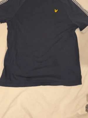 Blå t-shirt från Lyle & Scott - Mörkblå t-shirt från Lyle & Scott med korta ärmar och rutiga detaljer på axlarna. Klassisk gul logga på bröstet. Tillverkad i mjuk bomull för skön känsla. Perfekt till jeans eller shorts för en avslappnad look.